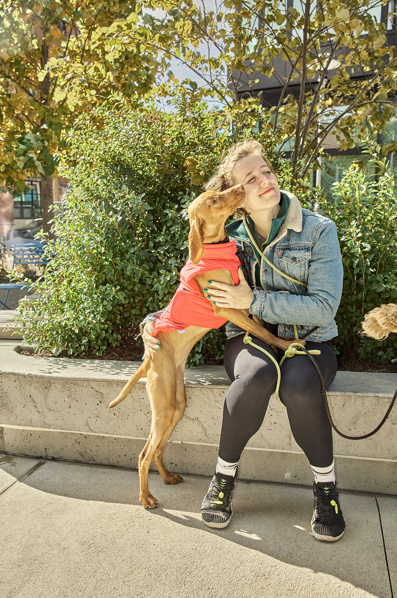 Jordan Phelps in a denim jacket with a small dog in a pink coat kissing her face