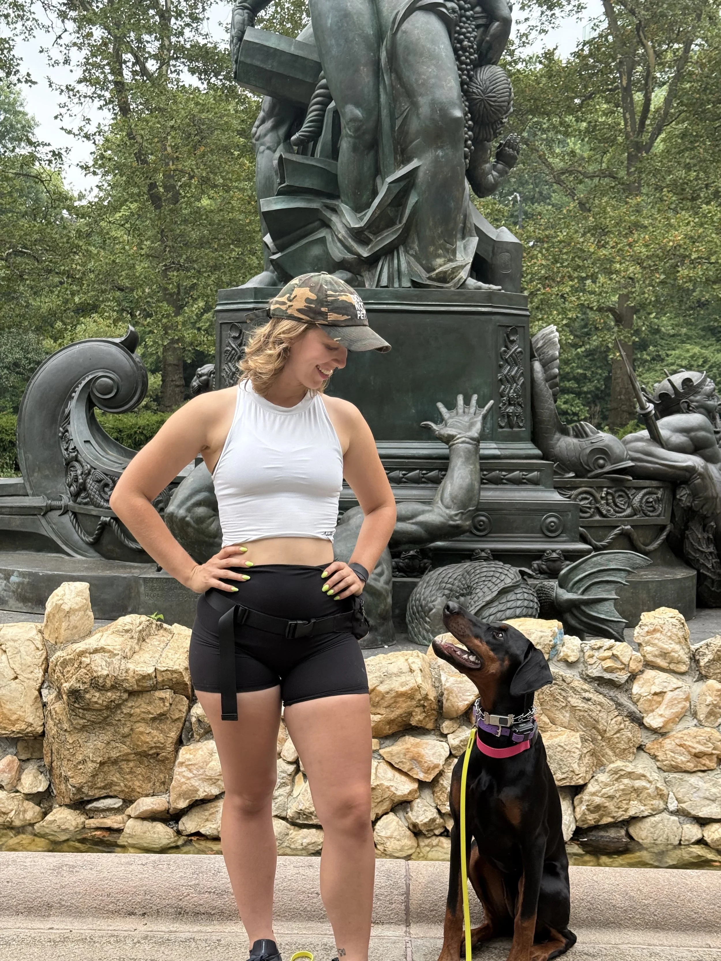 Jordan Phelps smiling beside a seated Doberman in front of a bronze monument in Brooklyn